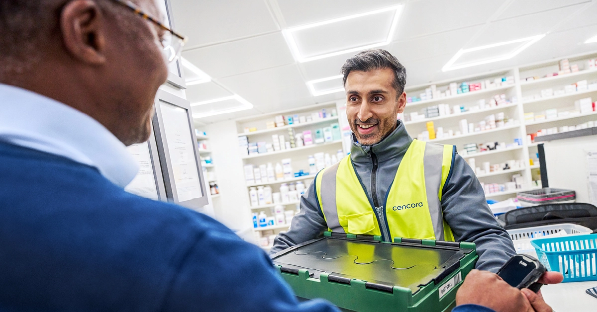 Pharmaceutical logistics worker wearing a high-visibility vest handing a secure green medical container to a healthcare professional inside a pharmacy.